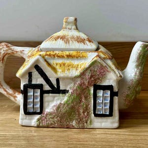 Puede incluir: Tetera de cerámica con forma de cabaña. El tejado es marrón y beige, las paredes son blancas con marcos de ventanas negros y un diseño floral verde y rosa. El asa y el pico son de color marrón claro y verde.