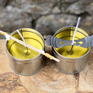 May include: Two silver metal candle tins with yellow wax and white wicks. One tin has a wooden wick holder, the other has a metal wick centering device. The tins sit on a wooden surface.