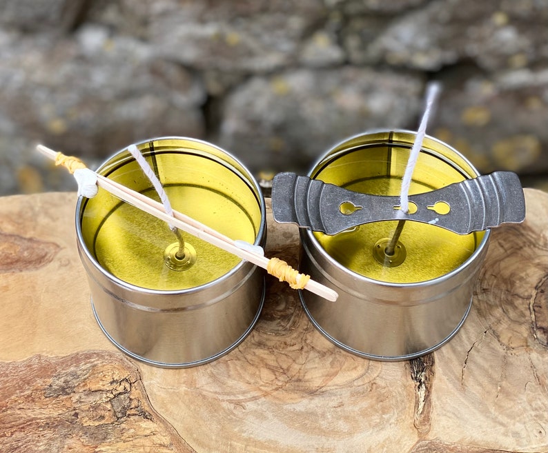 May include: Two silver metal candle tins with yellow wax and white wicks. One tin has a wooden wick holder, the other has a metal wick centering device. The tins sit on a wooden surface.