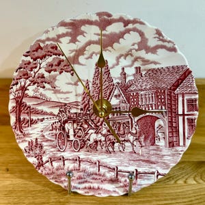 Op de afbeelding: Een decoratieve wandklok gemaakt van een bord met een rood en wit transferontwerp. De scène toont een paardenkoets op een weg, met een gebouw op de achtergrond. De wijzers van de klok zijn goudkleurig.