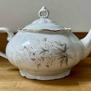 May include: A white porcelain teapot with a lid, handle, and spout. The teapot is decorated with a floral pattern of white flowers with red centers and grey leaves. The rim and spout have gold accents. The teapot is on a wooden surface.