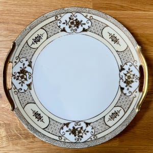 Puede incluir: Bandeja redonda para servir de color crema con ribetes y asas doradas. La bandeja presenta un centro blanco y está decorada con motivos florales y geométricos dorados alrededor del borde. Las asas también son doradas.