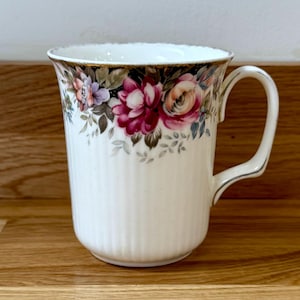 May include: White ceramic mug with a floral design featuring pink, red, and purple flowers. The rim is trimmed with gold, and the mug has a ribbed texture. The handle is white with a silver accent.