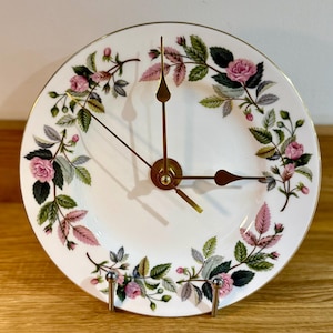 May include: A white ceramic plate clock with a gold rim and brass clock hands. The plate is decorated with pink roses, green leaves, and pink foliage. The clock is displayed on a metal stand.