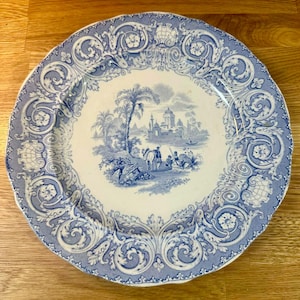 Puede incluir: Un plato redondo de estilo antiguo con un diseño azul y blanco. El centro presenta un paisaje con palmeras, figuras y edificios. El borde tiene un patrón detallado y ornamentado. El plato es de cerámica.