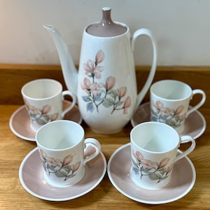 May include: A white ceramic coffee set with a floral design. The set includes a coffee pot with a brown lid, four cups, and saucers. The cups and coffee pot are decorated with pink and green floral patterns.