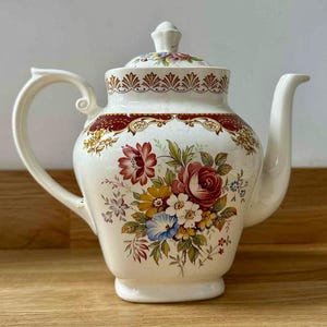 Pode incluir: Bule de cerâmica creme com um design floral. O bule tem uma alça curva, um bico e uma tampa com um pequeno botão. O design floral inclui flores vermelhas, rosa, amarelas e azuis. Uma borda bordô e dourada acentua a parte superior e inferior da tampa.