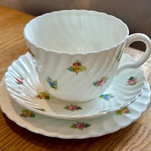 May include: A white teacup and saucer set with a floral pattern. The cup and saucer have a scalloped edge and are decorated with small pink, yellow, and blue flowers.