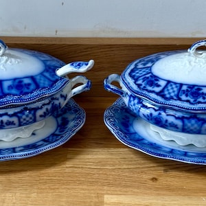 Op de afbeelding: Twee bijpassende witte keramische terrines met deksel en donkerblauwe bloemen- en ruitpatronen. Elke ovale terrine heeft zijhandgrepen, een decoratief deksel met een handvat en een bijpassende ovale serveerschaal met een geschulpte rand.