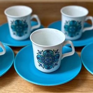 May include: A set of six white ceramic coffee cups with matching turquoise saucers. Each cup features a floral design in shades of teal and blue. The cups have a handle and are placed on a wooden surface.