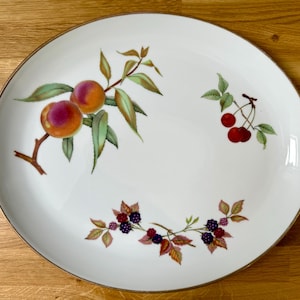 May include: White ceramic serving platter with a gold rim, decorated with illustrations of peaches, cherries, and blackberries. The fruit illustrations are in orange, red, purple, and green. The platter is on a wooden surface.