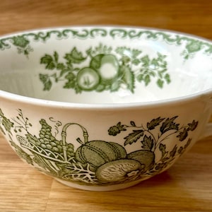 Może przedstawiać: Biała ceramiczna miska do zupy z dwoma uchwytami, z zielonym wzorem owoców i liści. Miska ma dekoracyjne obramowanie i szczegółową ilustrację winogron, melonów i innych owoców. Miska stoi na drewnianej powierzchni.