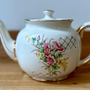 May include: Cream-colored ceramic teapot with a floral design featuring pink, red, and yellow flowers. The teapot has gold trim on the handle, spout, and lid. A decorative lattice pattern is also present.