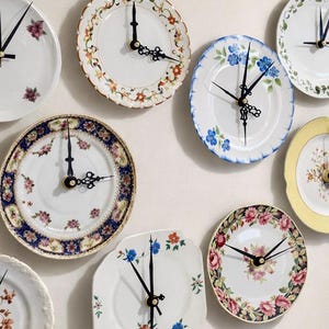 May include: A collection of decorative plate clocks with various floral patterns and designs. The clocks feature black hands and are mounted on a white wall. The plates have different shapes and colors, including white, blue, yellow, and floral patterns.