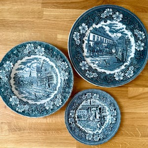 Pode incluir: Três pratos decorativos antigos azuis e brancos com bordas florais. Cada prato apresenta uma cena de paisagem detalhada no centro. Os pratos são dispostos sobre uma superfície de madeira, mostrando seus desenhos intrincados e charme vintage.