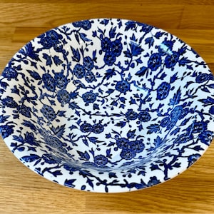 Könnte beinhalten: Eine weiße Keramikschüssel mit blauem Blumenmuster. Die Schüssel hat eine runde Form und ist mit einem sich wiederholenden Design aus blauen Blumen und Blättern verziert. Die Schüssel steht auf einer Holzoberfläche.
