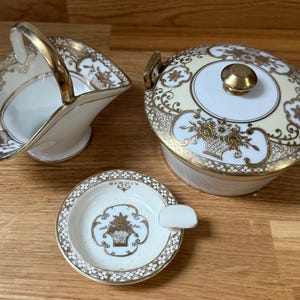 Könnte beinhalten: Dreiteiliges Vintage-Porzellan-Set mit goldenen Akzenten. Enthält einen Henkelkorb, eine Schüssel mit Deckel und einen kleinen Aschenbecher. Die Teile weisen florale und geometrische Muster auf cremefarbenem Hintergrund auf.