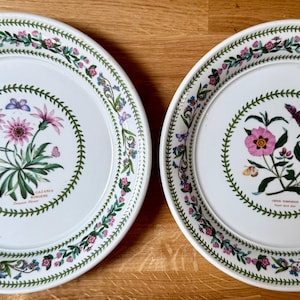 May include: Two white ceramic plates with floral designs. One plate features pink flowers and butterflies, the other has pink flowers. Both plates have a green border with floral and leaf patterns. The plates are on a wooden surface.