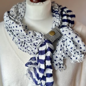 May include: A white and blue striped scarf with a white polka dot pattern and a gray leather scarf holder with a gold button.