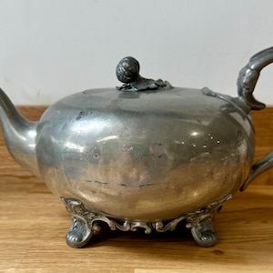 Puede incluir: Tetera antigua de color plateado con un pico curvo y un asa ornamentada. La tetera tiene un cuerpo redondeado y se apoya en tres patas decorativas. La tapa está rematada con un pequeño pomo redondo. Un clásico para los amantes del té.