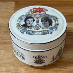 Könnte beinhalten: Eine runde, weiße Dose mit Deckel, die die Hochzeit von Prinz Charles und Lady Diana Spencer im Jahr 1981 zeigt. Der Deckel zeigt Porträts und Text, mit Illustrationen an den Seiten und einem goldenen Rand.