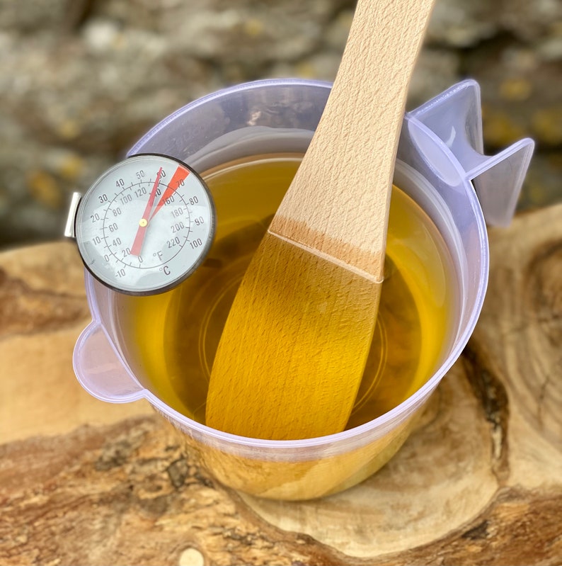 May include: A clear plastic measuring cup filled with a yellow liquid, a wooden spoon, and a thermometer. The thermometer reads approximately 160 degrees Fahrenheit. The spoon is partially submerged in the liquid.