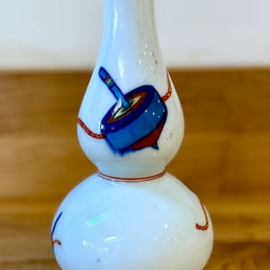 May include: White ceramic vase with a double-bulb shape, decorated with a blue and red spinning top design. The vase has red accents around the neck and base, with red rope-like details. A decorative piece for home decor.