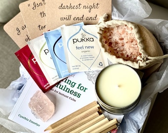 May include: A gift box filled with self-care items, including a book titled "Writing for Mindfulness", a pink crystal, a candle, a bag of pink Himalayan salt, tea bags, and colored pencils. The box also includes a card with the quote "Even the darkest night will end and..."