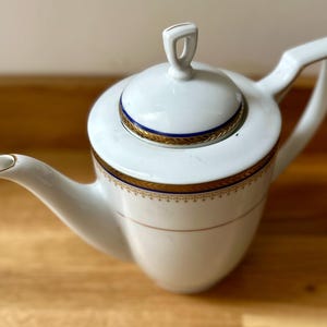Puede incluir: Tetera de porcelana blanca con tapa, con adornos dorados y azules. La tetera tiene un pico curvo y un asa con detalles dorados. El diseño es clásico y elegante, ideal para servir té.