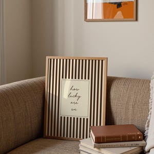 Op de afbeelding: Een ingelijste print met een licht houten frame. De print heeft verticale bruine en witte strepen met een centraal cr&egrave;mekleurig rechthoek. De rechthoek heeft de woorden "how lucky are we" in zwart schrift. Een stapel boeken staat voor de print.