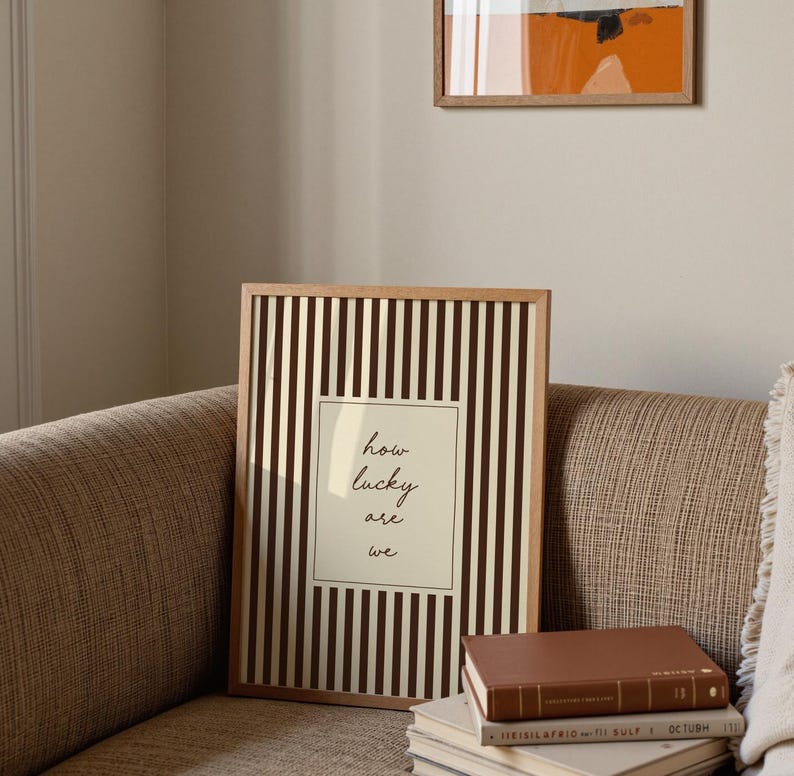 Op de afbeelding: Een ingelijste print met een licht houten frame. De print heeft verticale bruine en witte strepen met een centraal cr&egrave;mekleurig rechthoek. De rechthoek heeft de woorden "how lucky are we" in zwart schrift. Een stapel boeken staat voor de print.