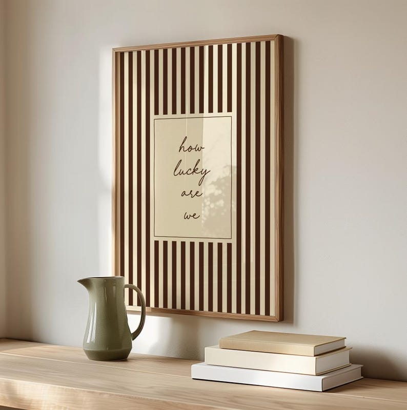 Op de afbeelding: Een ingelijst kunstwerk met een bruin en cr&egrave;mekleurig gestreept patroon en een centraal paneel met de woorden "how lucky are we" in zwart schrift. Een groene kan en gestapelde boeken staan op een houten oppervlak.