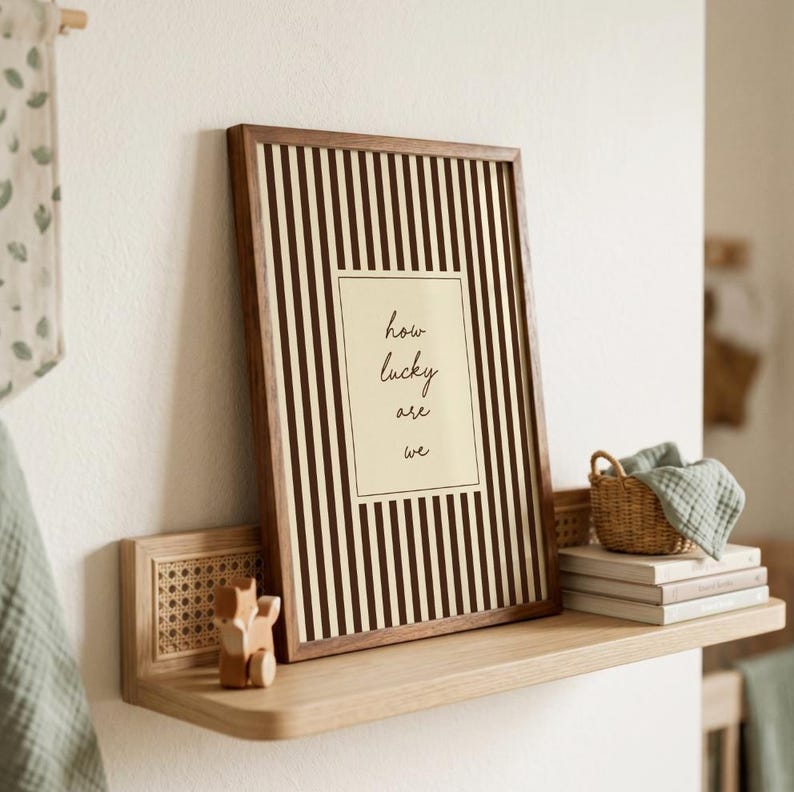 Op de afbeelding: Een ingelijste print met een bruine houten lijst en een gestreepte achtergrond. De print bevat de tekst 'how lucky are we' in handschriftstijl. De print wordt weergegeven op een houten plank met een klein mandje en boeken.