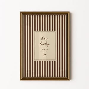 Op de afbeelding: Een ingelijste print met een goudkleurige lijst. De achtergrond heeft verticale bruine en cr&egrave;mekleurige strepen. Een cr&egrave;mekleurige rechthoek in het midden bevat de handgeschreven tekst "how lucky are we".