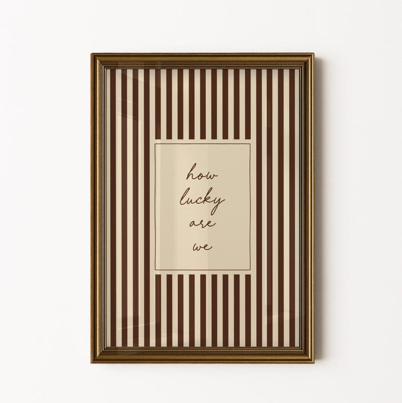 Op de afbeelding: Een ingelijste print met een goudkleurige lijst. De achtergrond heeft verticale bruine en cr&egrave;mekleurige strepen. Een cr&egrave;mekleurige rechthoek in het midden bevat de handgeschreven tekst "how lucky are we".