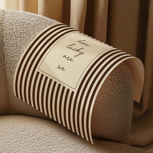 Op de afbeelding: Een beige en bruin gestreept decoratief papier is over een beige gestoffeerde armleuning gedrapeerd. Het papier bevat de tekst "how lucky are we" in een rechthoekige witte doos.