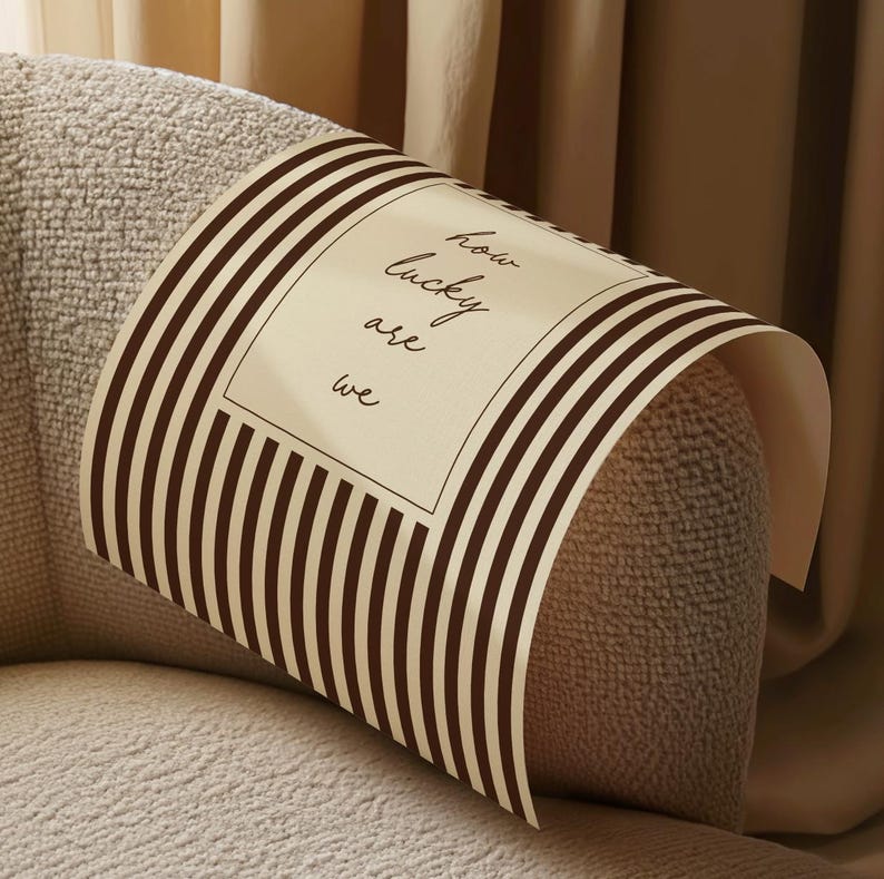 Op de afbeelding: Een beige en bruin gestreept decoratief papier is over een beige gestoffeerde armleuning gedrapeerd. Het papier bevat de tekst "how lucky are we" in een rechthoekige witte doos.