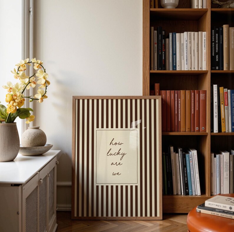 Op de afbeelding: Een ingelijste kunstprint met een verticaal gestreept patroon in bruin en cr&egrave;me. De print toont de tekst "how lucky are we" in een handschriftstijl. Het kunstwerk wordt tentoongesteld in de buurt van een boekenkast en decoratieve vazen.