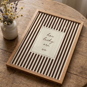 Op de afbeelding: Een ingelijste print met een bruin en wit gestreepte achtergrond en de tekst "how lucky are we" in een rechthoekige doos. De lijst is gemaakt van lichtgekleurd hout. Een kleine vaas met bloemen staat op de achtergrond.