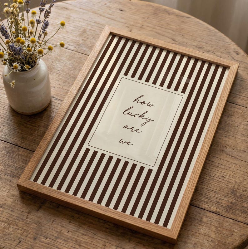 Op de afbeelding: Een ingelijste print met een bruin en wit gestreepte achtergrond en de tekst "how lucky are we" in een rechthoekige doos. De lijst is gemaakt van lichtgekleurd hout. Een kleine vaas met bloemen staat op de achtergrond.