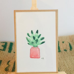 Peut inclure: Une peinture à l'aquarelle d'une plante verte dans un pot rose. La peinture est sur un petit chevalet en bois.