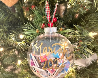 May include: A clear glass Christmas bauble with a gold inscription that reads "Mariah 2022". The bauble is filled with small pieces of paper with writing on them.