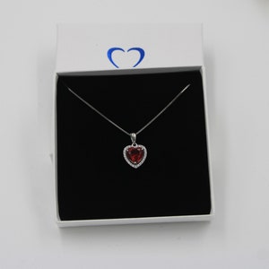 Könnte beinhalten: Halskette mit herzförmigem Anhänger, rotem Edelstein in der Mitte und einem Kranz aus kleineren, klaren Steinen. Der Anhänger hängt an einer filigranen Silberkette, präsentiert in einer weißen Geschenkbox mit schwarzem Innenraum.
