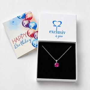 Könnte beinhalten: Eine silberne Halskette mit einem runden rosa Edelsteinanhänger in einer weißen Geschenkbox mit dem blauen Schriftzug "exclusiv 4 you". Daneben liegt eine Grußkarte mit bunten Aquarellballons und dem Text "HAPPY Birthday" in Blau und Rot.