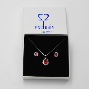 Könnte beinhalten: Ein silbernes Halsketten- und Ohrring-Set mit einem roten ovalen Edelstein-Anhänger, der von kleineren klaren Edelsteinen umgeben ist. Der Schmuck befindet sich in einer weißen Schachtel mit dem Text "Ω2 exclusiv 4 you".