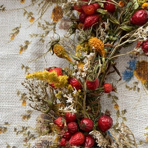 May include: A dried floral wreath with red rose hips, yellow dried flowers, and white dried flowers. The wreath is on a white floral patterned fabric background.