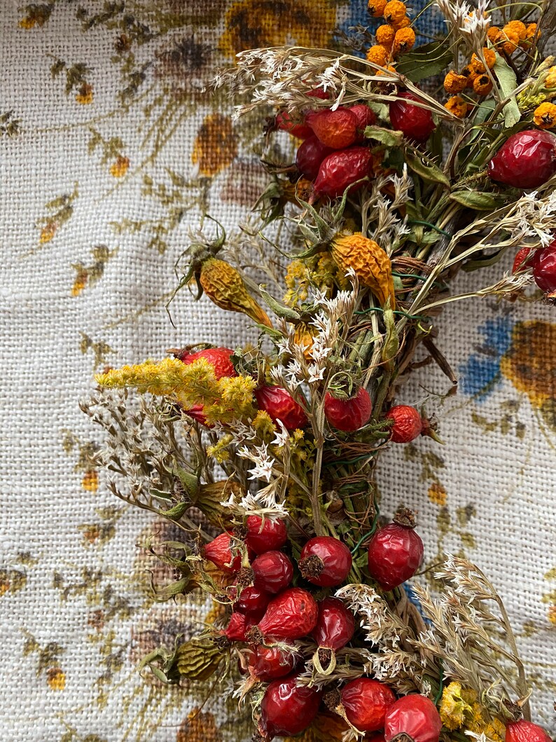 May include: A dried floral wreath with red rose hips, yellow dried flowers, and white dried flowers. The wreath is on a white floral patterned fabric background.