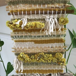 K&ouml;nnte beinhalten: Ein gewebter Wandbehang mit einem nat&uuml;rlichen Holzrahmen und einer Vielzahl von Texturen, darunter Stroh, Garn und getrocknete Blumen. Der Wandbehang hat eine neutrale Farbpalette in Beige, Gelb und Gr&uuml;n.