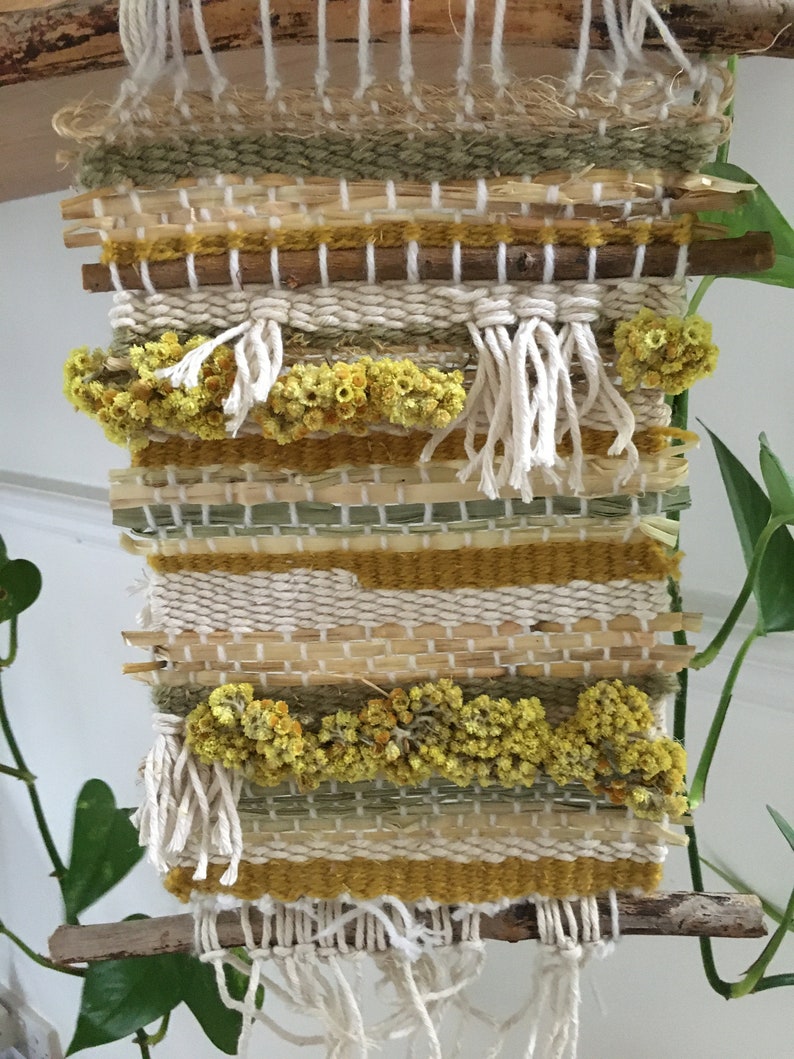 K&ouml;nnte beinhalten: Ein gewebter Wandbehang mit einem nat&uuml;rlichen Holzrahmen und einer Vielzahl von Texturen, darunter Stroh, Garn und getrocknete Blumen. Der Wandbehang hat eine neutrale Farbpalette in Beige, Gelb und Gr&uuml;n.