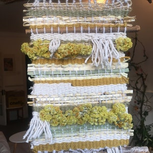 K&ouml;nnte beinhalten: Ein gewebter Wandbehang mit Naturfasern und getrockneten Blumen. Der Behang ist aus verschiedenen Texturen und Farben gefertigt, darunter Wei&szlig;, Beige, Gr&uuml;n und Gelb. Der Behang ist an zwei Holzst&ouml;cken befestigt.